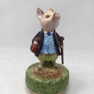 May include: A ceramic figurine of a pig dressed in a blue vest, black jacket, and green pants, holding a brown bag and a cane. The pig stands on a green grassy base with a light beige bottom.
