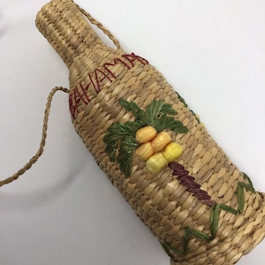 Può includere: Borsa a forma di bottiglia in paglia intrecciata, con tracolla. Presenta un disegno di palma con frutta gialla e arancione e foglie verdi. La parola "BAHAMA" è ricamata con filo rosso.