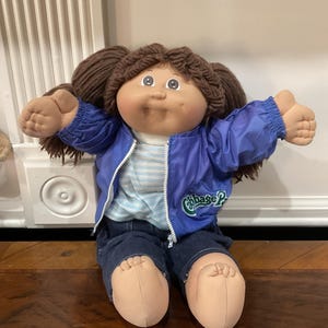 May include: A Cabbage Patch Kids doll with brown yarn hair styled in pigtails, wearing a blue zip-up jacket with the Cabbage Patch logo, a light blue and white striped shirt, and dark blue denim pants. The doll has a soft fabric body and limbs.