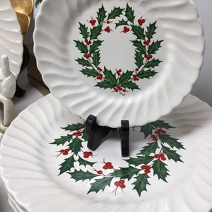 May include: White ceramic plates with a swirled edge design. Each plate features a Christmas wreath design with green leaves and red berries. The plates are stacked, with one plate displayed on a black stand.