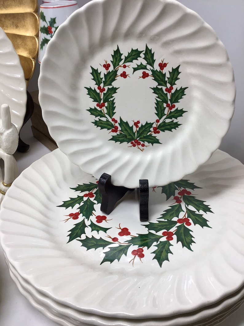 May include: White ceramic plates with a swirled edge design. Each plate features a Christmas wreath design with green leaves and red berries. The plates are stacked, with one plate displayed on a black stand.