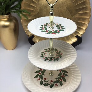 May include: A three-tiered serving tray with a gold handle and stand. Each white plate features a holly wreath design with red berries and green leaves. The plates have a ridged edge.