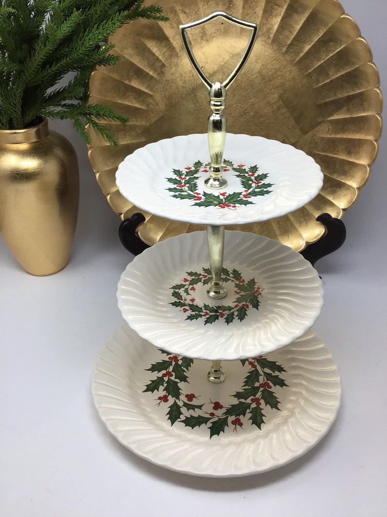 May include: A three-tiered serving tray with a gold handle and stand. Each white plate features a holly wreath design with red berries and green leaves. The plates have a ridged edge.