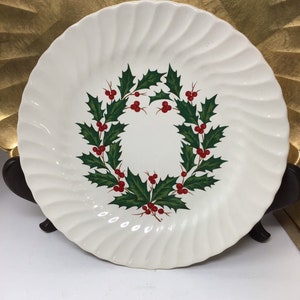May include: A white ceramic plate with a wavy rim design. The plate features a wreath of green holly leaves and red berries in the center. The plate is likely for serving food and is a festive holiday item.