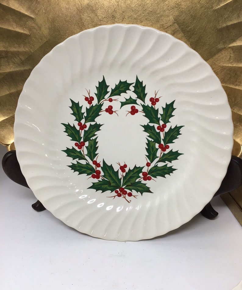 May include: A white ceramic plate with a wavy rim design. The plate features a wreath of green holly leaves and red berries in the center. The plate is likely for serving food and is a festive holiday item.