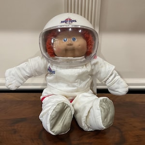 Puede incluir: Muñeca Cabbage Patch Kids vestida de astronauta. La muñeca tiene un traje espacial blanco con detalles rojos, un casco blanco con visera transparente y pelo rojo. Está sentada sobre una superficie de madera.