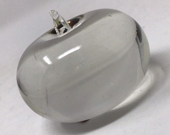 May include: A clear glass apple sculpture with a small stem. The apple is smooth and rounded, with a slight indentation where the stem is attached. The glass is transparent, allowing light to pass through.