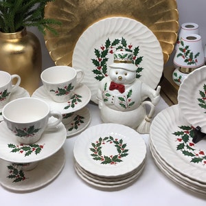 May include: A collection of white ceramic tableware with a Christmas holly design. The set includes cups, saucers, plates, and a snowman-shaped serving piece. The plates have a ridged edge. A gold vase and decorative plate are in the background.