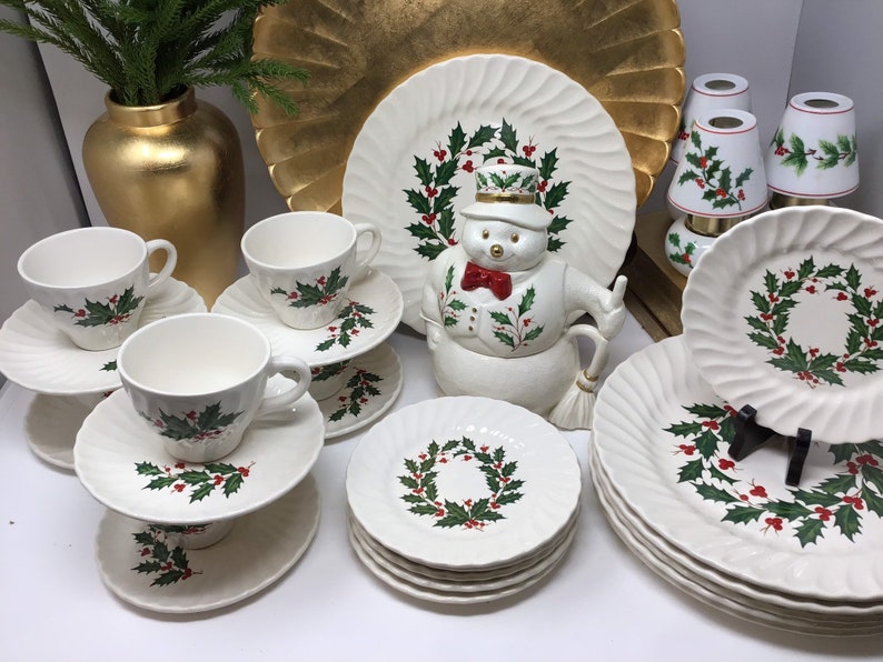 May include: A collection of white ceramic tableware with a Christmas holly design. The set includes cups, saucers, plates, and a snowman-shaped serving piece. The plates have a ridged edge. A gold vase and decorative plate are in the background.