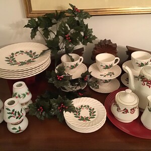 May include: A complete Christmas-themed tea set with plates, cups, saucers, a teapot, creamer, and sugar bowl. The white porcelain pieces feature a holly wreath design and red ribbon accents. The set includes several small lamp shades with holly designs.