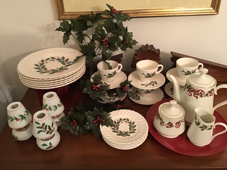 May include: A complete Christmas-themed tea set with plates, cups, saucers, a teapot, creamer, and sugar bowl. The white porcelain pieces feature a holly wreath design and red ribbon accents. The set includes several small lamp shades with holly designs.