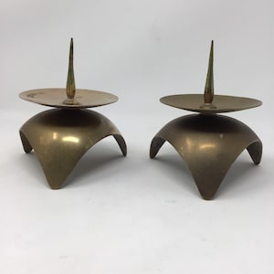 May include: Two vintage brass candle holders. Each has a rounded base with three curved legs, a flat circular plate, and a central spike for holding a candle. The brass has a warm, aged patina, suggesting they are antique or vintage.