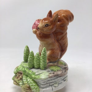 Puede incluir: Figura de cerámica de una ardilla marrón con una cola tupida, sosteniendo un objeto rosa. Está posada sobre una formación rocosa con árboles verdes. La base es de color beige claro. Objeto decorativo.