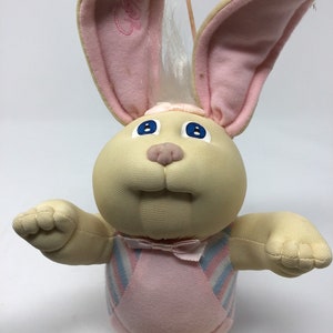 May include: A vintage plush bunny doll with a cream-colored face and body, large pink-lined ears, and blue eyes. The doll wears a pink and blue striped outfit and a small bow tie. The bunny has a soft, cuddly appearance.