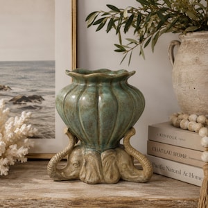 May include: A decorative ceramic vase featuring a teal, ribbed, scalloped-edge bowl supported by a base sculpted into the likeness of three elephant heads with trunks extending outward. The vase has a glossy finish and sits on a flat surface.