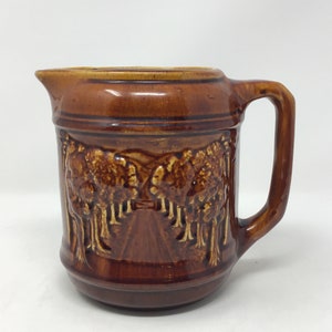 May include: A brown ceramic jug with a handle and spout. The jug features a raised relief design of a road lined with trees. The colour is a rich, glossy brown with lighter brown accents.