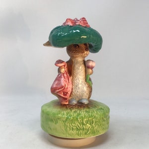Peut inclure: Figurine en céramique représentant un lapin portant un chapeau vert avec un nœud rouge, tenant un sac rose et une fleur. Le lapin est posé sur une base verte et herbeuse. Objet décoratif.
