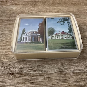 May include: A deck of playing cards in a clear plastic case. The cards feature images of historic buildings. One card shows a white building with columns, and the other shows a white building with a red roof. The case is beige.