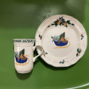 May include: A white ceramic mug and plate set with a Christmas theme. The mug and plate feature a blue sleigh design with a Christmas tree and gifts. The rims are decorated with green and gold accents.