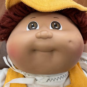 May include: A doll with a round vinyl face, brown yarn hair, and brown eyes. The doll wears a yellow corduroy hat and matching yellow corduroy overalls over a white shirt with a white ribbon tie at the neck. The doll has a dimple on the left cheek.