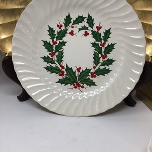May include: White ceramic plate with a spiral edge design. The plate features a wreath of green holly leaves and red berries in the center. The plate is displayed on a dark brown stand, perfect for holiday dining and decor.