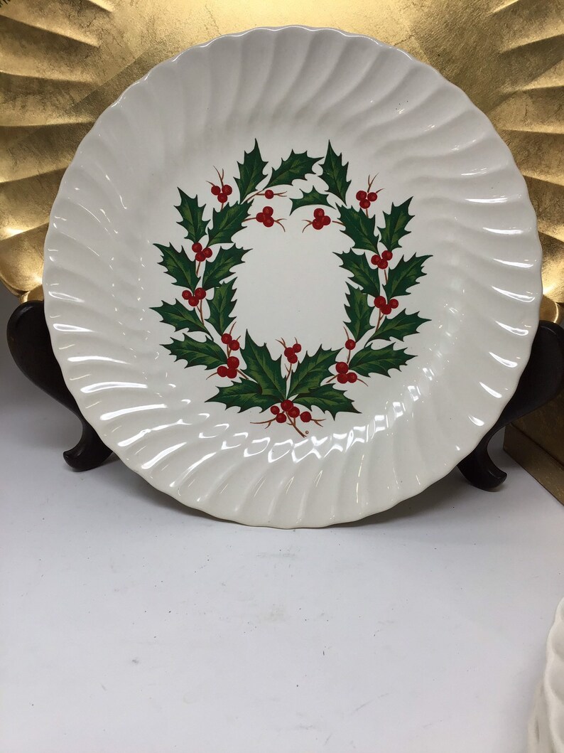 May include: White ceramic plate with a spiral edge design. The plate features a wreath of green holly leaves and red berries in the center. The plate is displayed on a dark brown stand, perfect for holiday dining and decor.