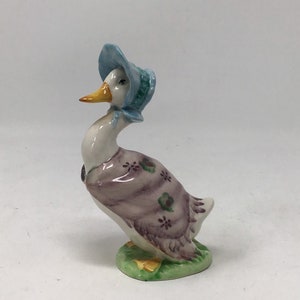 May include: A ceramic goose figurine, wearing a blue bonnet and a lavender dress with floral detailing. The goose stands on a green base with yellow accents. It has a white body, a yellow beak, and black eyes.