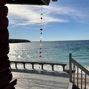 May include: A vertical hanging mobile composed of a series of colorful sea glass pieces and small dark stones strung together on a thin wire, suspended from the edge of a wooden porch roof overlooking a body of water.