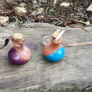 May include: Two small, handmade ceramic bottles with cork stoppers and leather or ribbon accents. One is purple and pink, the other is blue and orange. They sit on a weathered wooden surface.