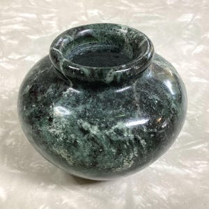 May include: A small, round, polished stone vase featuring a deep forest green color with intricate white and dark grey veining throughout. The vessel has a smooth, glossy finish, a wide, rounded body, and a thick, circular opening at the top.