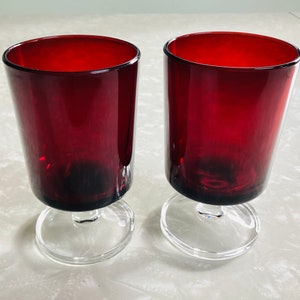 May include: Two red glass tumblers with clear glass stems and bases. The glasses are empty and sitting on a white surface.
