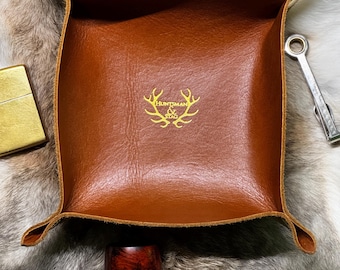 May include: A brown leather tray with the words "Huntsman & Stag" in gold lettering. A pipe, lighter, and wrench are next to the tray. The tray is on a fur-like surface.