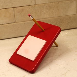 May include: A rectangular red wooden stand angled upward, featuring a square white notepad attached to its surface and a circular hole near the top. A yellow wooden pencil with a pink eraser is inserted through the hole in the red stand.