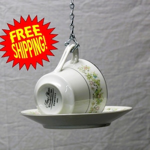 May include: A white Fine China teacup and saucer set, suspended by a silver chain. The cup is decorated with floral patterns in green, yellow, and pink, and the saucer has a matching design. The set is labeled "Romance."