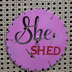 May include: A circular, pink metal sign with the words "She Shed" in silver and red. The sign has a saw blade edge and is mounted on a perforated metal surface.