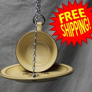 May include: A hanging bird feeder made from a cream-colored teacup and saucer set. The teacup and saucer are suspended by a silver chain. The saucer has a brown floral design. The teacup has a brown rim.
