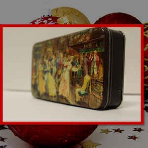 May include: A vintage tin box with a colorful illustration of people dancing in a tavern. The tin box is brown with a black lid and is surrounded by red and gold Christmas ornaments.