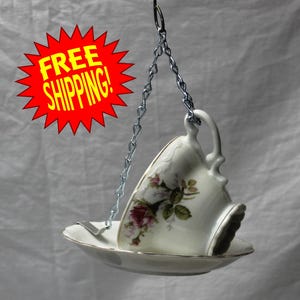 May include: A vintage teacup bird feeder, suspended by a silver chain. The white teacup and saucer feature a floral design with pink and green accents. The teacup is upside down, creating a unique bird feeder.