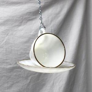 May include: A white teacup and saucer set with gold trim, suspended by a silver chain. The teacup is round with a delicate handle, and the saucer is scalloped. The set is against a soft, white background.