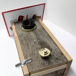 May include: A wooden box with a dark gray stain and a light wood trim. The box has a black plastic lid with a gold metal band around it. The lid has the words "Turn on Top" printed on it. The box also has a small silver cross and a gold metal knob.