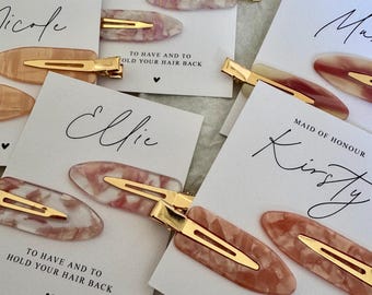 Puede incluir: Varias horquillas para el cabello con cierres dorados se exhiben en tarjetas blancas. Las horquillas tienen un patrón veteado en tonos rosa y blanco. Las tarjetas tienen nombres escritos como "Bella" y "Ellie", y el texto "TO HAVE AND TO HOLD YOUR HAIR BACK".