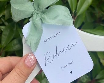 Puede incluir: Percha blanca con un lazo de cinta verde salvia y una tarjeta blanca adjunta. La tarjeta dice "BRIDESMAID" y "Rebecca" en escritura cursiva. La fecha "24.08.2023" y un pequeño corazón están impresos debajo del nombre.