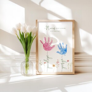 May include: A framed print with a light wood frame displays two handprints, one pink and one blue, with the names "Penelope" and "George" below. The text "If Nanas were flowers, we'd pick you!" is at the top. A vase of white tulips is to the left.