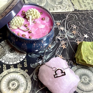 May include: Pink candle with dried flowers and pink crystals in a blue and gold tin with a lid. A silver chain necklace with a Libra charm is on a pink rose quartz crystal. A green soap bar is on a black and white patterned surface.