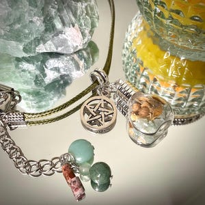 May include: A collection of mystical jewelry and accessories. Includes a silver pentagram pendant, a glass bulb pendant filled with dried herbs, and a chain bracelet with gemstone charms. The background features a green crystal and a decorative glass.