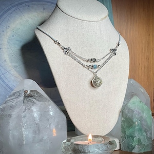 May include: A silver-toned necklace with three strands, displayed on a mannequin. The necklace features decorative floral accents, small blue beads, and a round pendant. The necklace is displayed with clear quartz crystals and a lit candle in a glass holder.