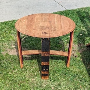 May include: A small, round wooden table made from reclaimed oak barrel staves. The tabletop is a light brown color, and the legs are a darker brown with red accents. The table is approximately 24 inches in diameter and stands about 20 inches tall.