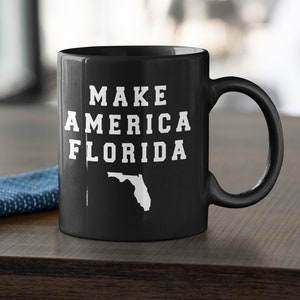 May include: Black ceramic mug with the text "MAKE AMERICA FLORIDA" in white block letters, along with a white outline of the state of Florida. The mug has a curved handle and is set on a wooden surface.