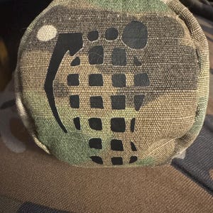 May include: A circular pouch in a camouflage pattern, featuring a black grenade design. The pouch has a loop at the top and is made from a robust, textured material. The colours include shades of green, brown, and tan.