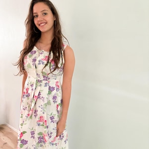 May include: A white sleeveless midi dress with a floral print featuring pink, purple, and green flowers. The dress has a button-up front and a fitted bodice.